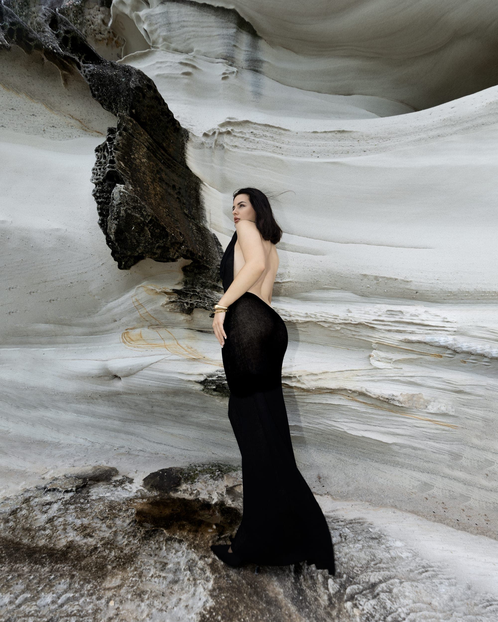 Fashion model in a black gown, side profile against layered sandstone rock, Sydney coastline photoshoot